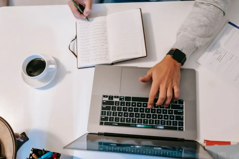A person working at a laptop at home, taking notes in a journal beside a cup of coffee